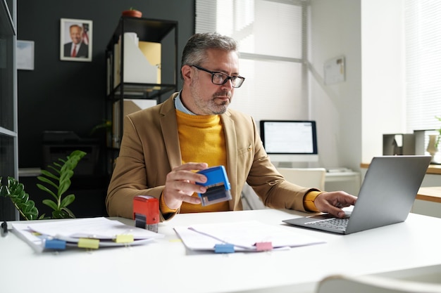 A person sitting at a desk, reviewing a budget on a laptop while holding a credit card. The background is a modern office with blurred figures moving around, suggesting a busy workplace.